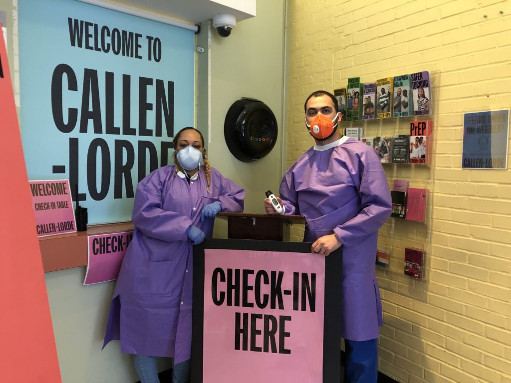 Two members of Callen-Lorde's staff in purple and blue PPE waiting at check-in stand to check patients' temperatures as they arrive for their appointments. 